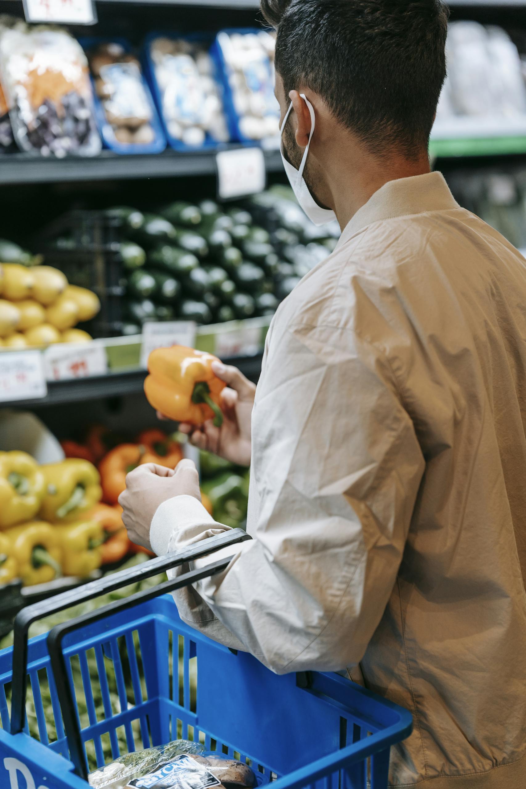 Back view of anonymous male buyer in protective mask picking ripe bell pepper while standing in vegetable department in supermarket